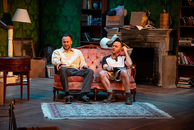 two men sit on a couch in a set resembling a living room.